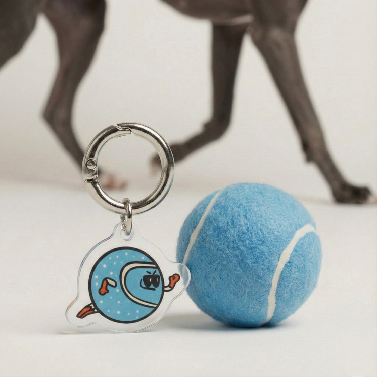 Fur Sie The Fetcher charm next to a blue tennis ball. The acrylic charm features a matching blue color and round ball-inspired shape