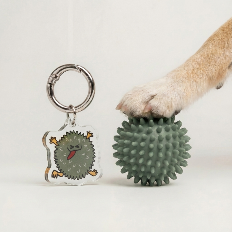 Fur Sie Troublemaker charm next to a green spiky rubber dog ball toy under a dog's paw. The acrylic charm mimics the toy's spiky texture and green color