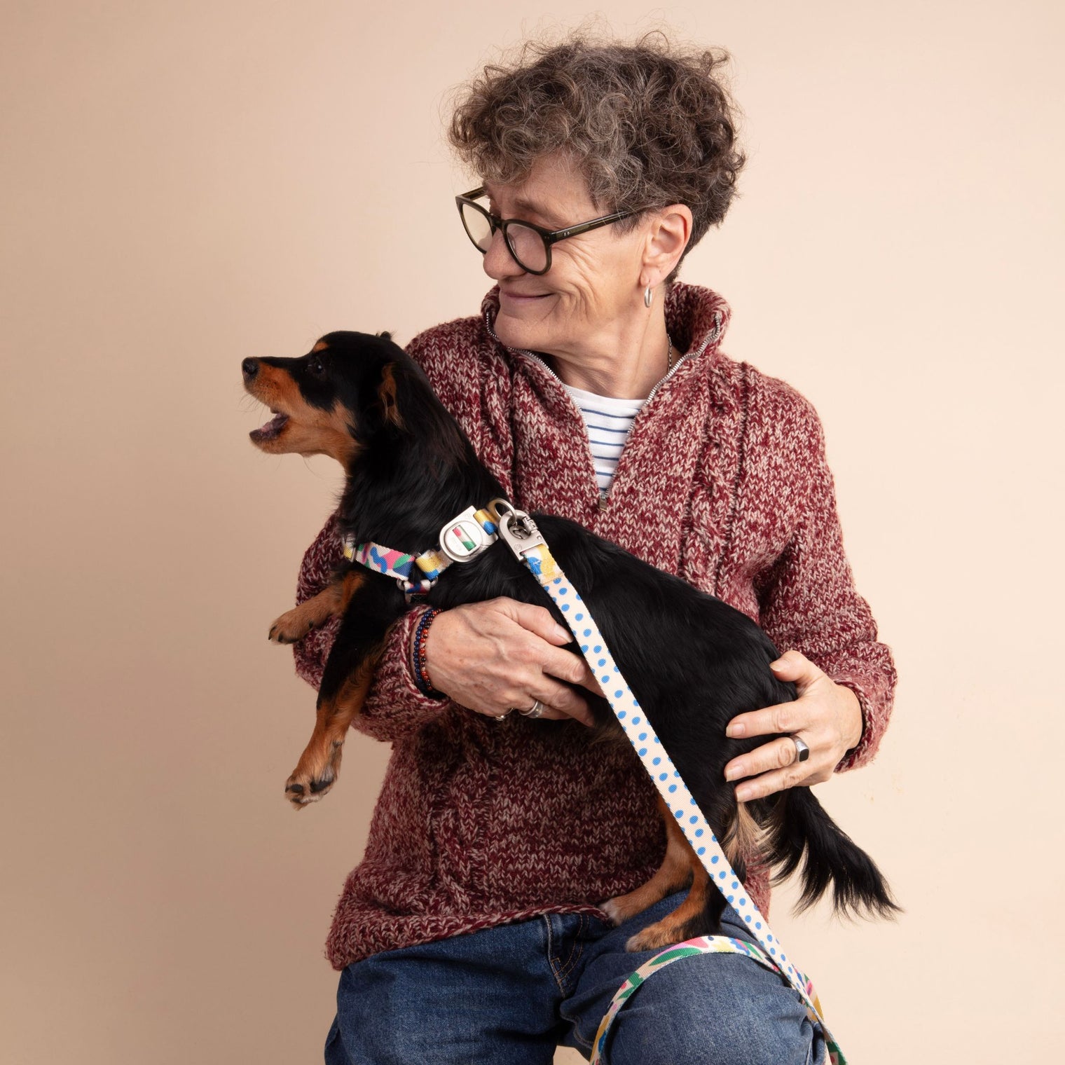 Woman holding a small black dog with a colorful Fur Sie Gaston harness and leash against a beige background