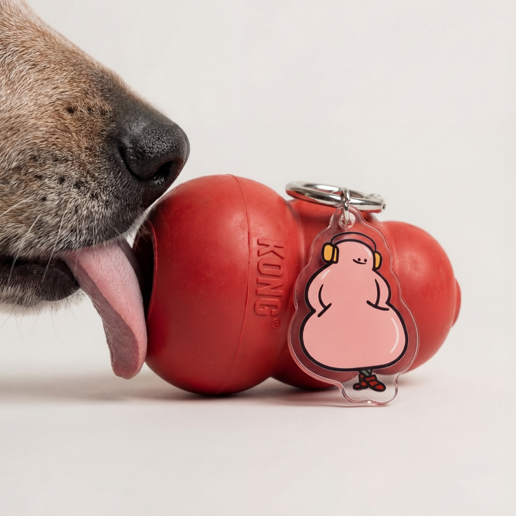A Fur Sie Pleaser charm next to a large red classic KONG chew toy being licked by a dog. The pink acrylic charm mimics the toy’s tiered shape and features a character with yellow headphones and red sneakers