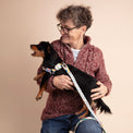 Woman holding a small black dog with a colorful Fur Sie Gaston harness and leash against a beige background